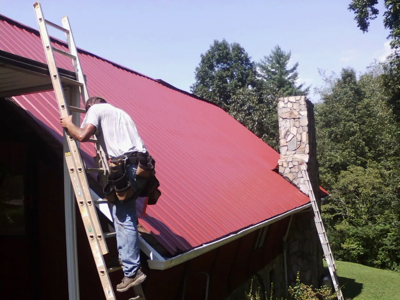 Roofer on a ladder beside a red metal roof and stone chimney — Soffit & Fascia Repair by Roofing Brothers in Brainerd